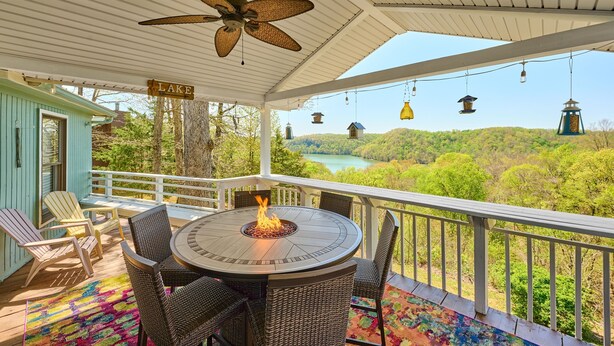 A covered outdoor deck with a fire pit table, wicker chairs, and a view of a lake and forest. - Renovated Cottage! Overlooks Dale Hollow! (Byrdstown)