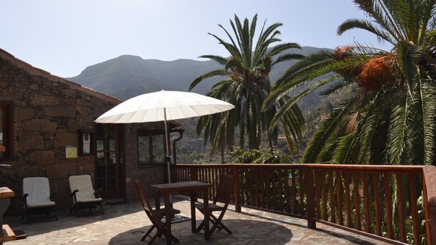 Un bâtiment en pierre doté d’un patio comprenant une table et des chaises, un parasol, et offrant une vue sur des palmiers et des montagnes.