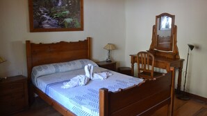 Un lit bien fait, recouvert d’une couvre-lit à motifs, et agrémenté d’une paire de serviettes blanches. On aperçoit une tête de lit en bois et une petite table de chevet avec une lampe. Un grand tableau encadré représentant une scène de forêt est accroché au mur au-dessus du lit.