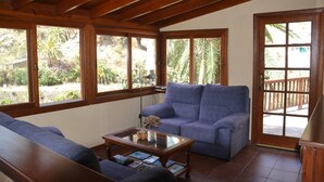 Un salon chaleureux avec un canapé bleu, une table basse en verre et des poutres en bois au plafond. De grandes fenêtres offrent une vue sur la verdure environnante.