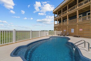 A swimming pool with a wooden deck, a white fence, and a building with balconies. - Premier Soundfront Dog Friendly w/ Pool, Hot Tub, Windsurfing Access, Theater (Rodanthe)