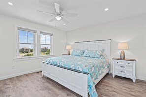 A bedroom with a bed, two bedside tables, a ceiling fan, and a window with a view of houses. - Premier Soundfront Dog Friendly w/ Pool, Hot Tub, Windsurfing Access, Theater (Rodanthe)