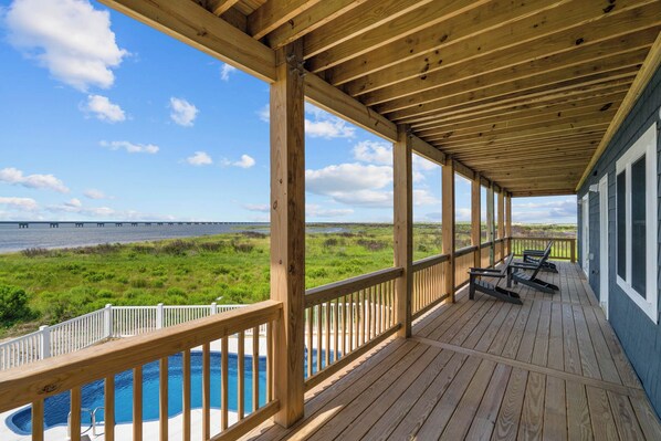 A wooden deck with a pool, overlooking a grassy area and a body of water with a bridge in the distance. - Premier Soundfront Dog Friendly w/ Pool, Hot Tub, Windsurfing Access, Theater (Rodanthe)