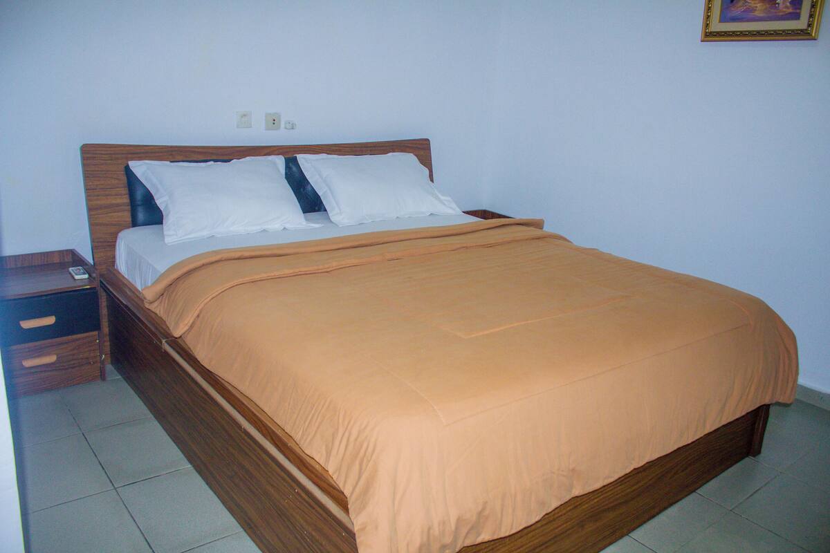 A single bed with a wooden headboard, white pillows, and an orange bedspread in a room with white walls and tiled flooring.