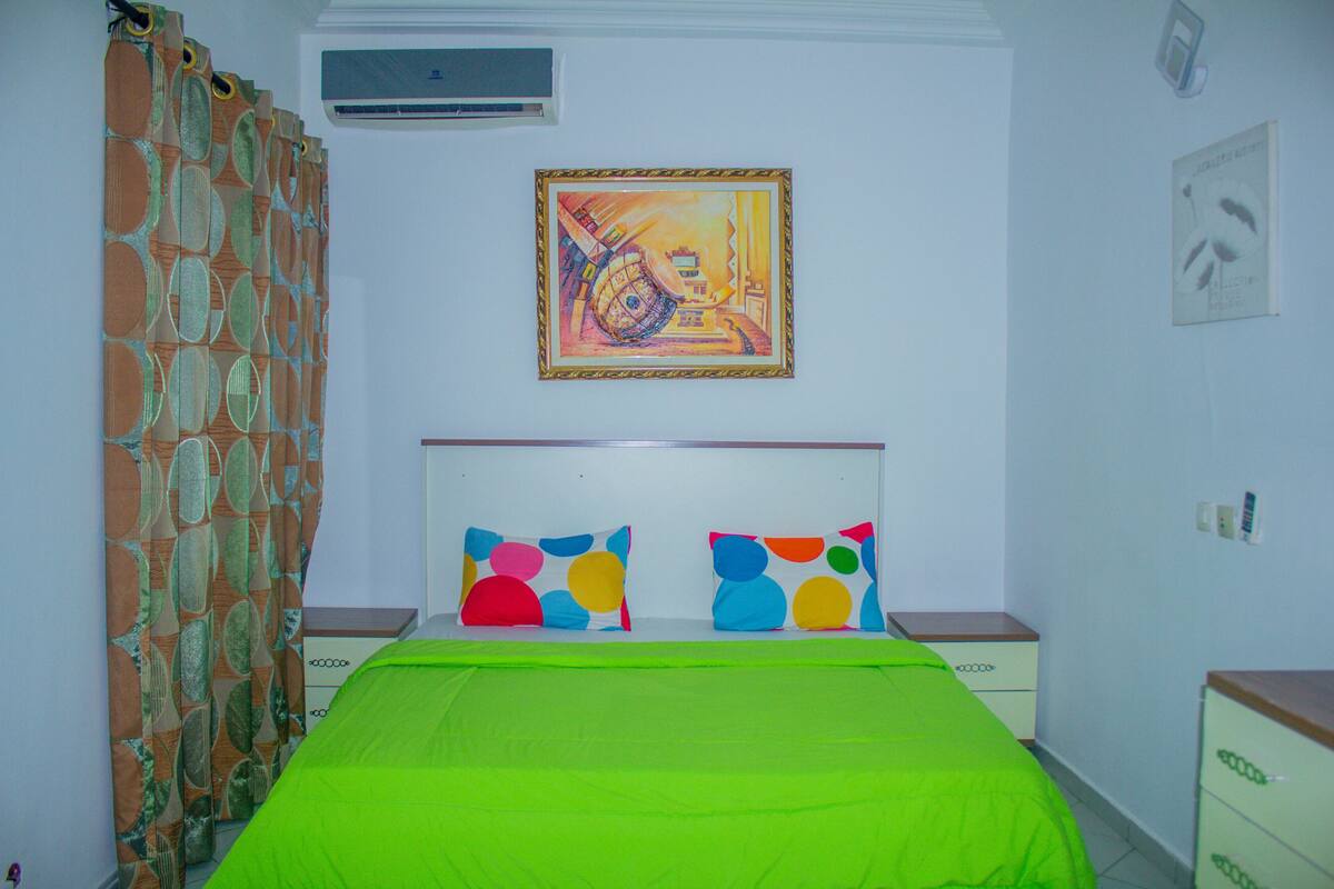 A bedroom with a green bedspread, a patterned curtain, and a colorful painting on the wall.