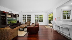A modern living room with a flat-screen TV, a brown leather sofa, wicker chairs, and a dining area with white bar stools. - Clanalpine Retreat  Leura (Leura)