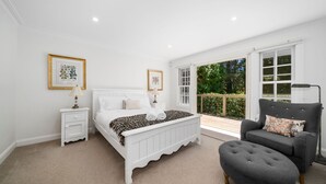 A bedroom with a white bed, two bedside tables, a grey armchair, and a large window with a view of greenery. - Clanalpine Retreat  Leura (Leura)