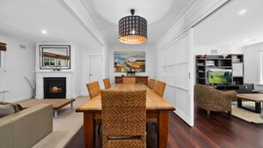 A modern living room with a fireplace, a dining table with woven chairs, a large painting, and a television in a dark entertainment unit. - Clanalpine Retreat  Leura (Leura)