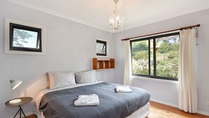 Un dormitori amb un llit gran, una taula de nit amb una làmpada, un penjallum, una finestra amb cortines i una estanteria sobre el llit.