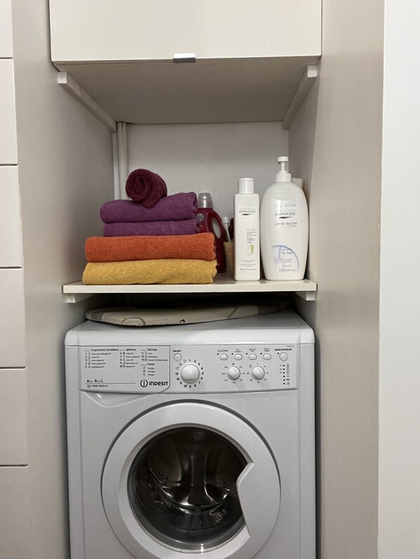 Une machine à laver dotée d’un panneau de commande et d’une étagère au-dessus, où sont rangées des serviettes pliées et des articles de toilette.