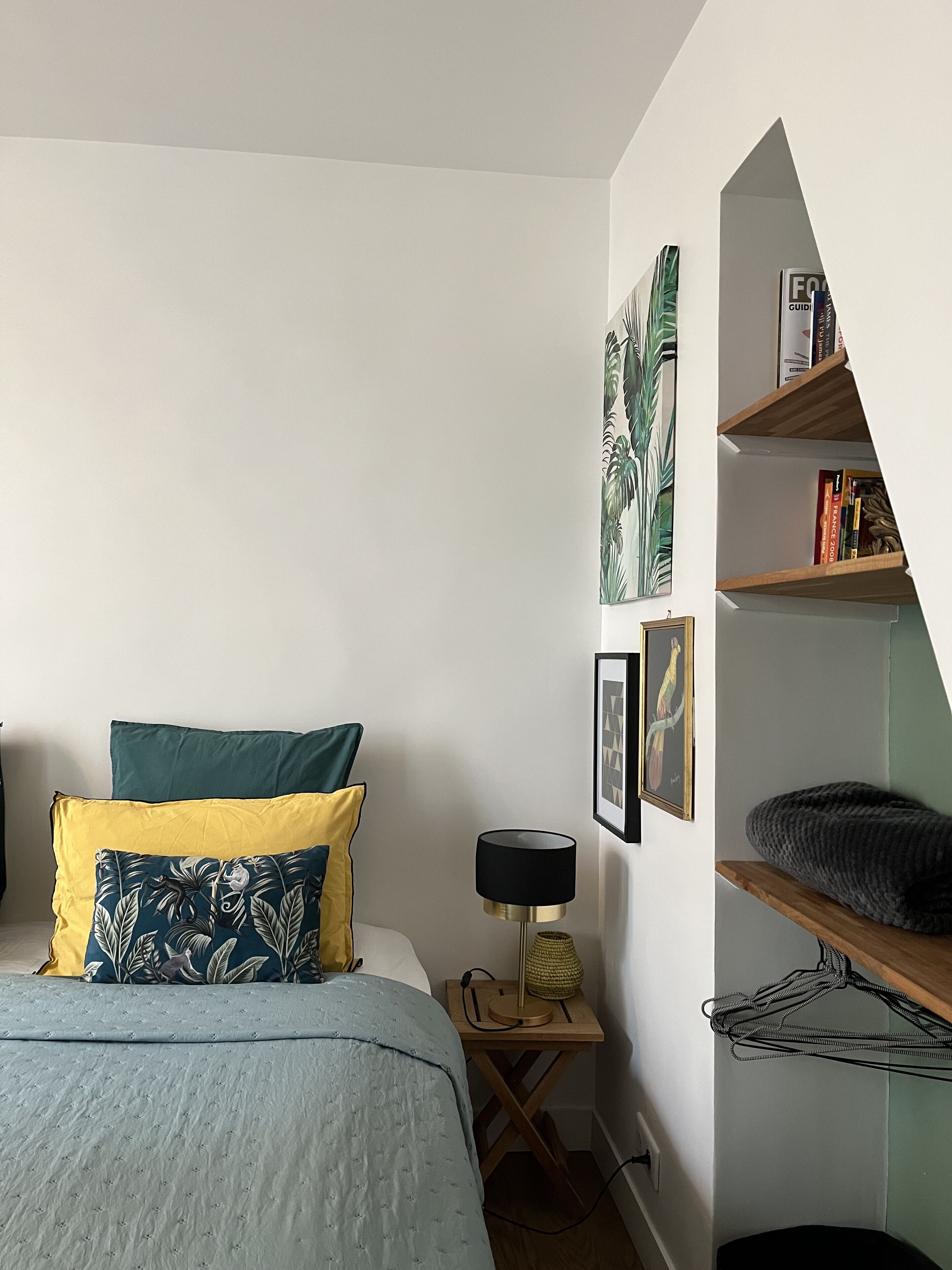 Ein Schlafzimmer mit einem Bett, Kissen, einem Nachttisch, einer Lampe, einem gerahmten Bild und einem Bücherregal.