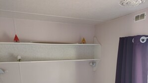 A small, single-bed room with a headboard, a shelf above the bed, and a window with a curtain.