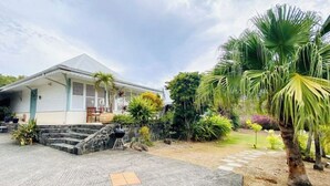 A house with a covered porch, a stone staircase, and a garden with various plants and a palm tree. - Villa Gaïa: Bungalow Ti-zen & spa - Parc Tropical (LE DIAMANT)