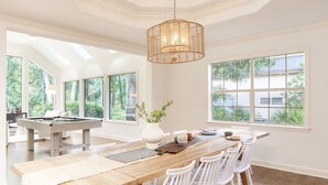 A spacious dining area with a long wooden table, white chairs, and a central chandelier. - Hilton Head Retreat • Private Pool + Deck Dining + Near Beach | Hilton Lagoon (Hilton Head Island)