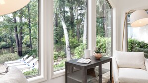 A room with large windows offering a view of a forested area, a white armchair, a small table with a vase, and a lamp. - Hilton Head Retreat • Private Pool + Deck Dining + Near Beach | Hilton Lagoon (Hilton Head Island)