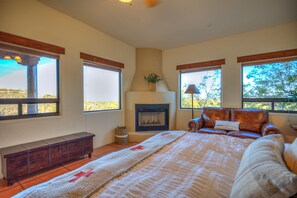 Une chambre avec une cheminée, un grand lit, un fauteuil en cuir et une vue sur l’extérieur grâce à de grandes fenêtres.
