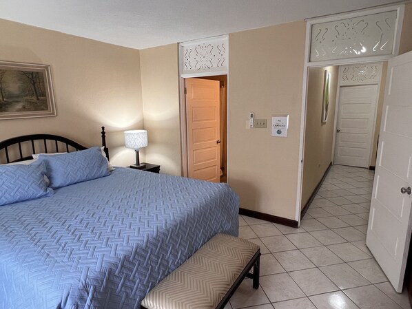Un dormitorio con cama, lámpara de noche, banca, una puerta e interruptor de luz en la pared.