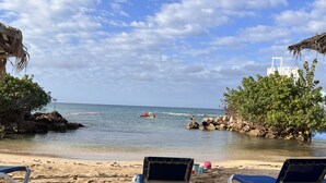 Una spiaggia con due lettini bianchi e azzurri, una costa rocciosa e persone che fanno kayak in acqua.