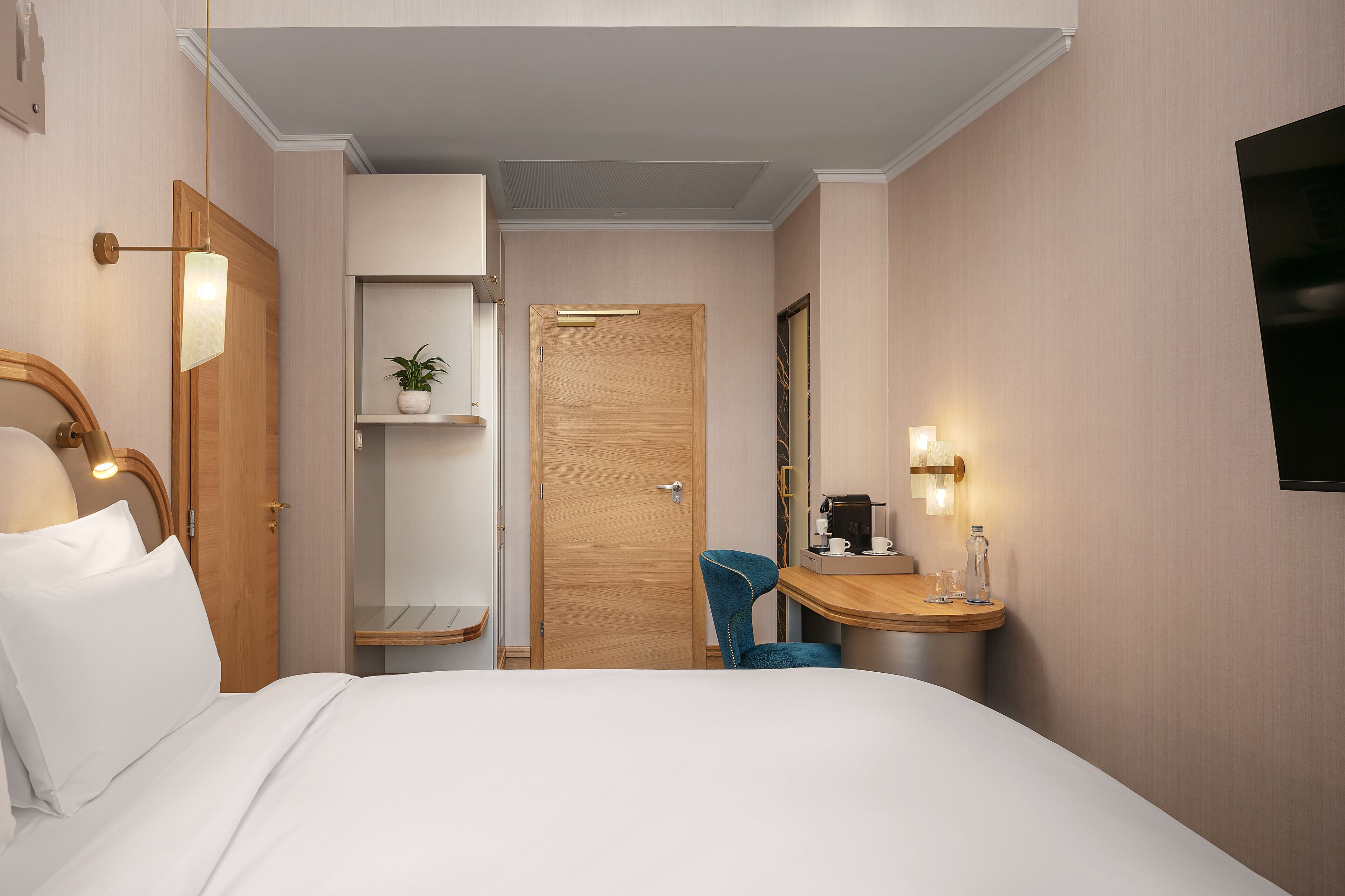 A hotel room with a bed, a desk with a chair, a shelf with a plant, and a door.