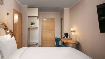 A hotel room with a bed, a desk with a chair, a shelf with a plant, and a door.