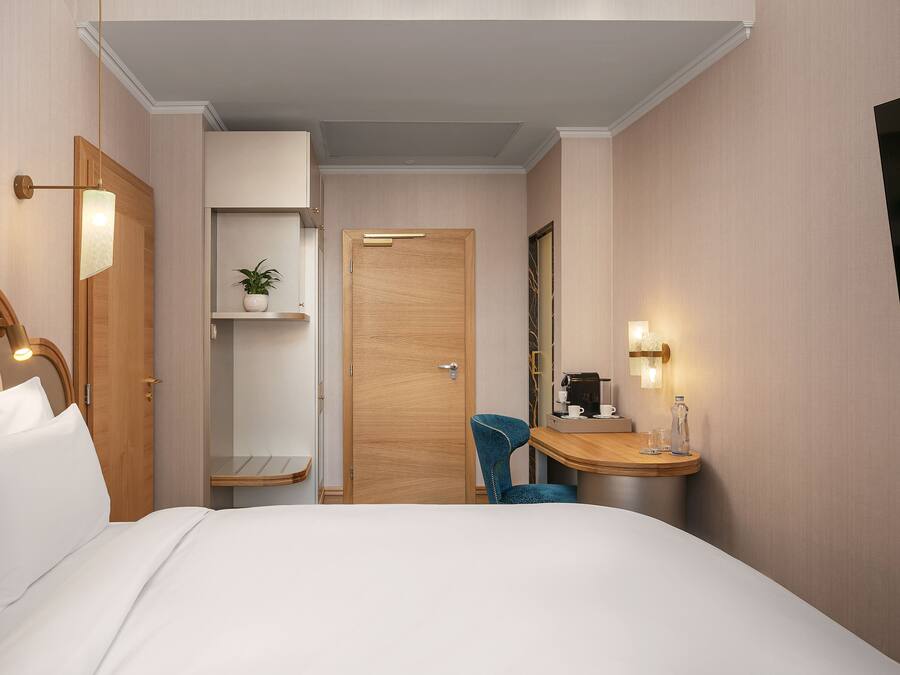 A hotel room with a bed, a desk with a chair, a shelf with a plant, and a door.