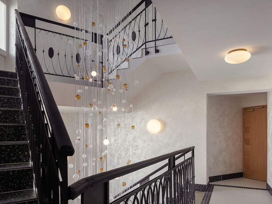 Una escalera con barandilla negra, una pared con luces colgantes y una puerta de madera al final.