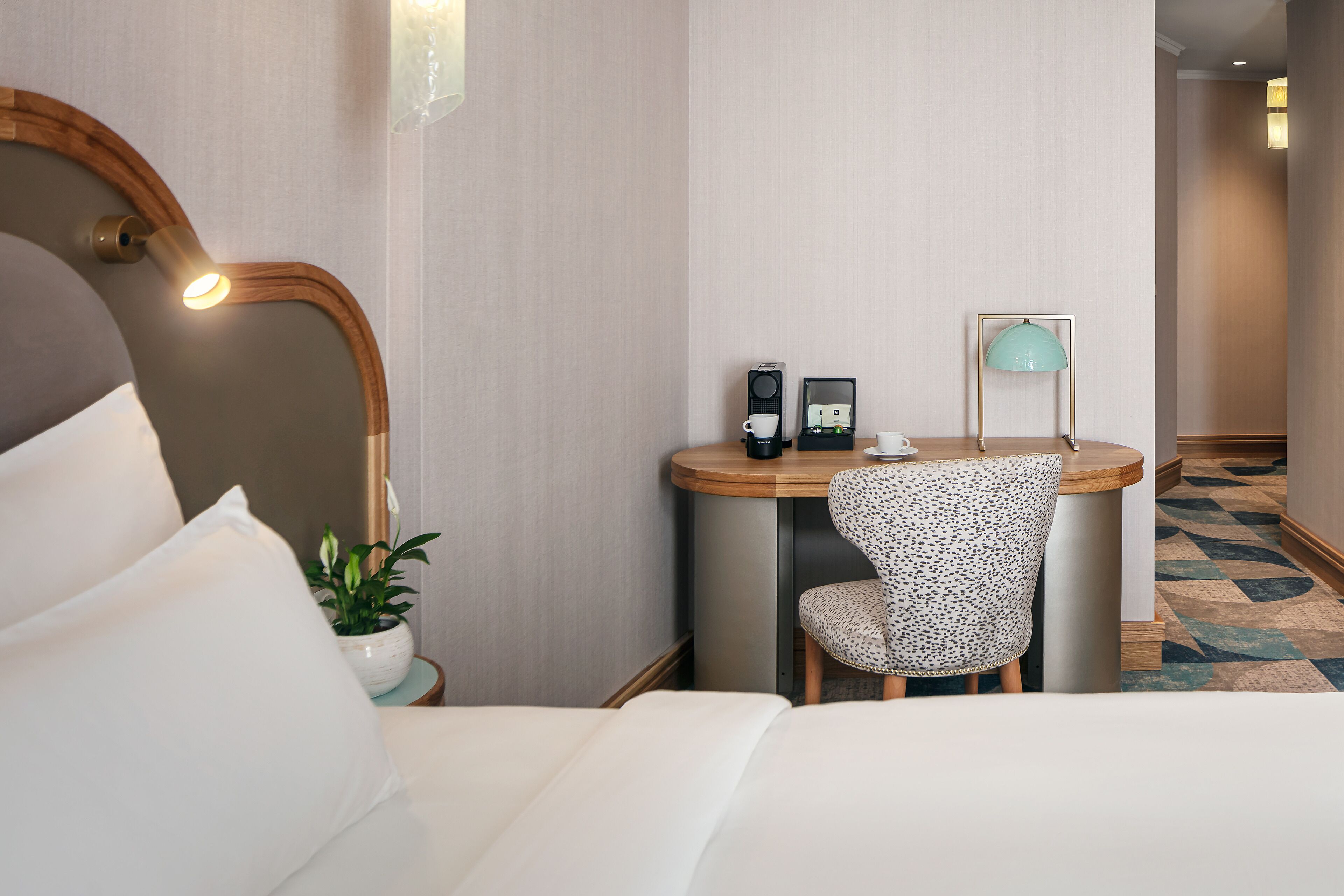 A hotel room with a bed, a desk with a chair, a mirror, a small table with a lamp, and a potted plant.