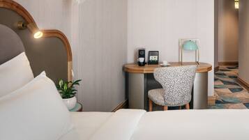 A hotel room with a bed, a desk with a chair, a mirror, a small table with a lamp, and a potted plant.