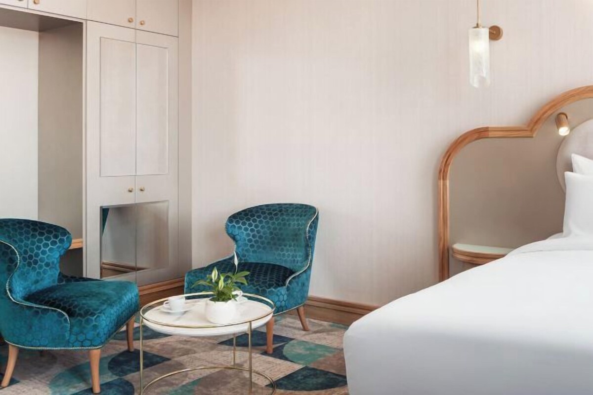 A hotel room with a bed, two blue upholstered chairs, a small round table with a plant, a white cabinet, and a mirror.