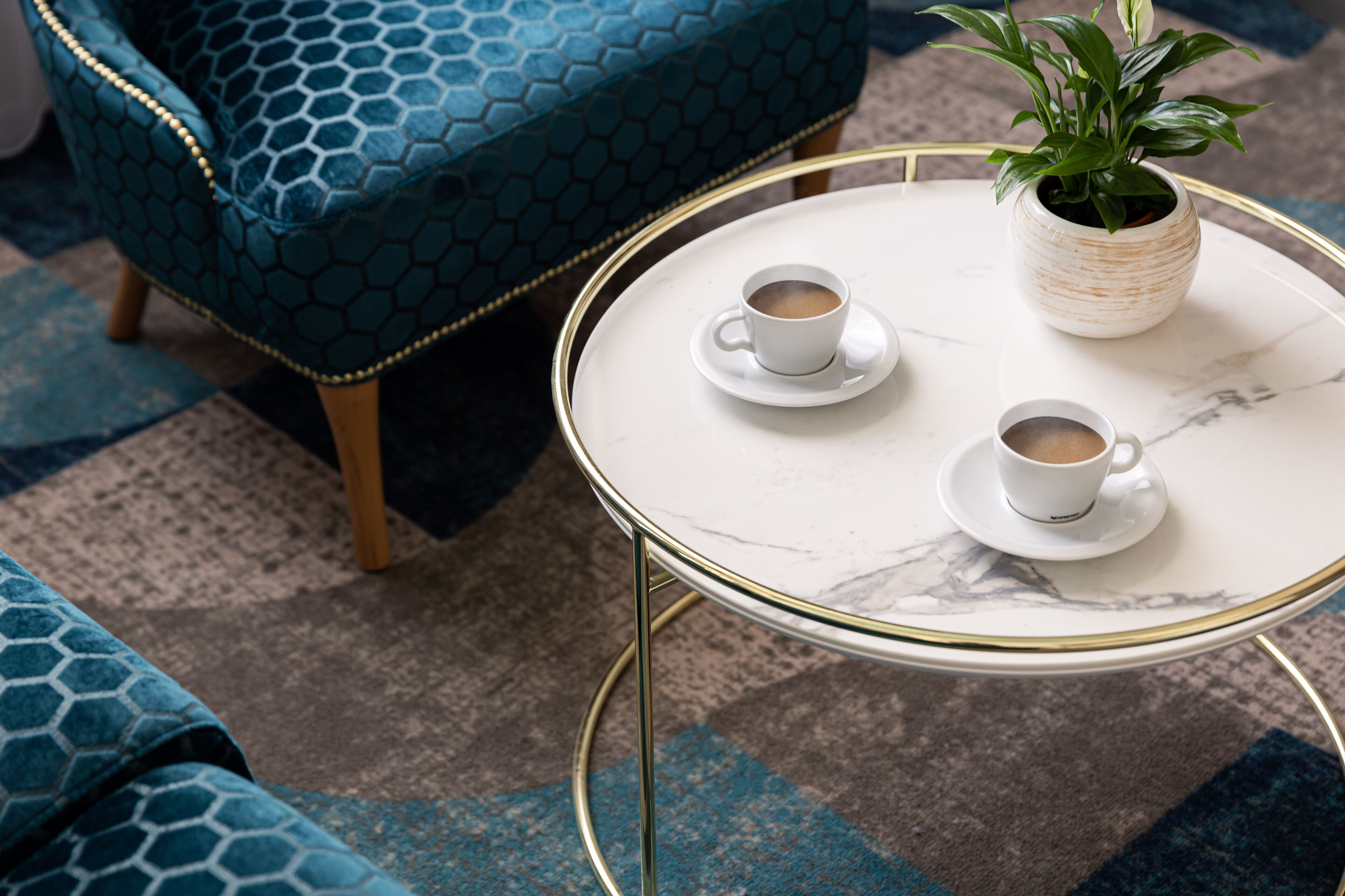 A round table with two white cups of coffee, a potted plant, and a blue patterned chair.