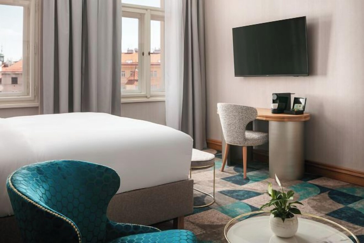 A hotel room with a bed, a blue armchair, a small table with a plant, a TV on the wall, and a view of buildings outside.