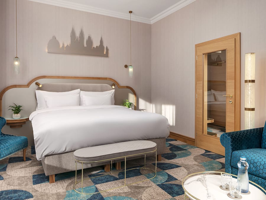 Habitación de hotel con una cama grande, dos sillones tapizados en azul, una mesita redonda con una botella y vasos, una mesita de noche con una planta, y una puerta de madera con un espejo.