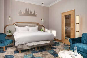 Quarto de hotel com uma cama grande, duas poltronas azuis estofadas, uma mesa redonda pequena com uma garrafa e copos, uma mesa de cabeceira com uma planta e uma porta de madeira com um espelho.