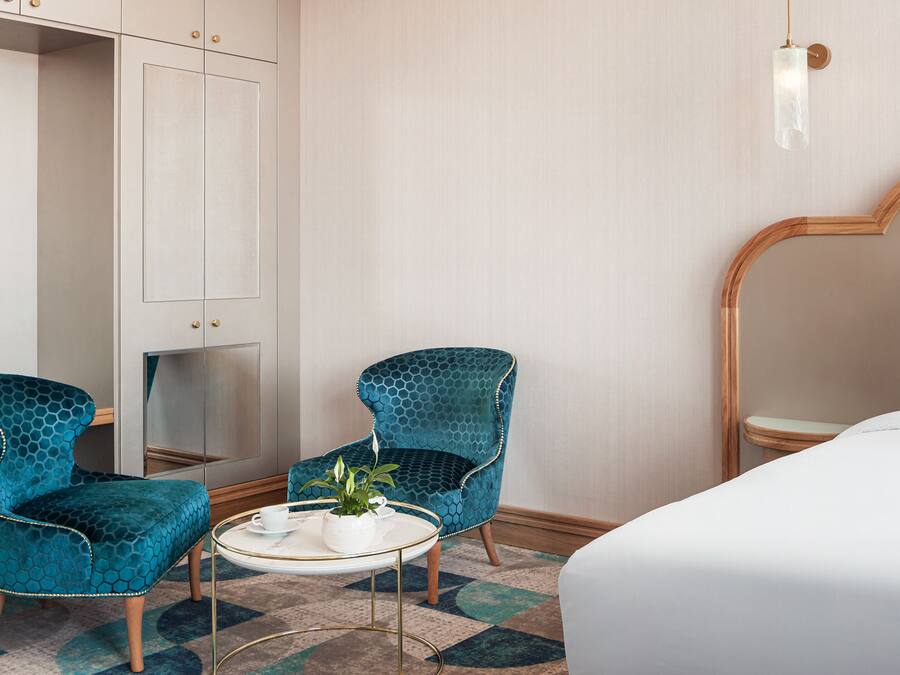 Habitación de hotel con una cama, dos sillas, una mesita con una planta y un espejo en la pared.