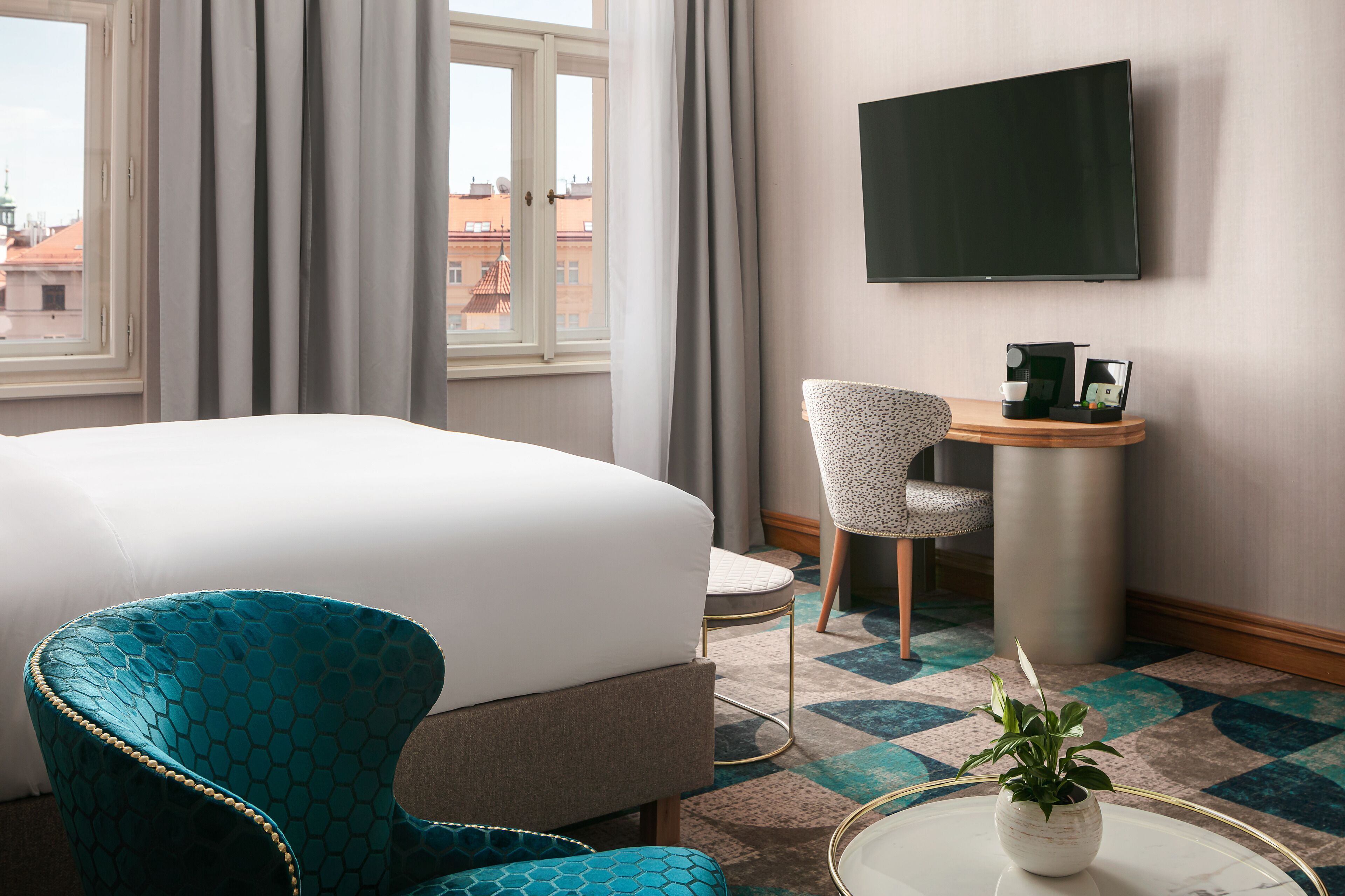 Ein Hotelzimmer mit einem Bett, einem blauen Sessel, einem kleinen Tisch mit Pflanze, einem Schreibtisch mit Stuhl und einem Fernseher an der Wand.