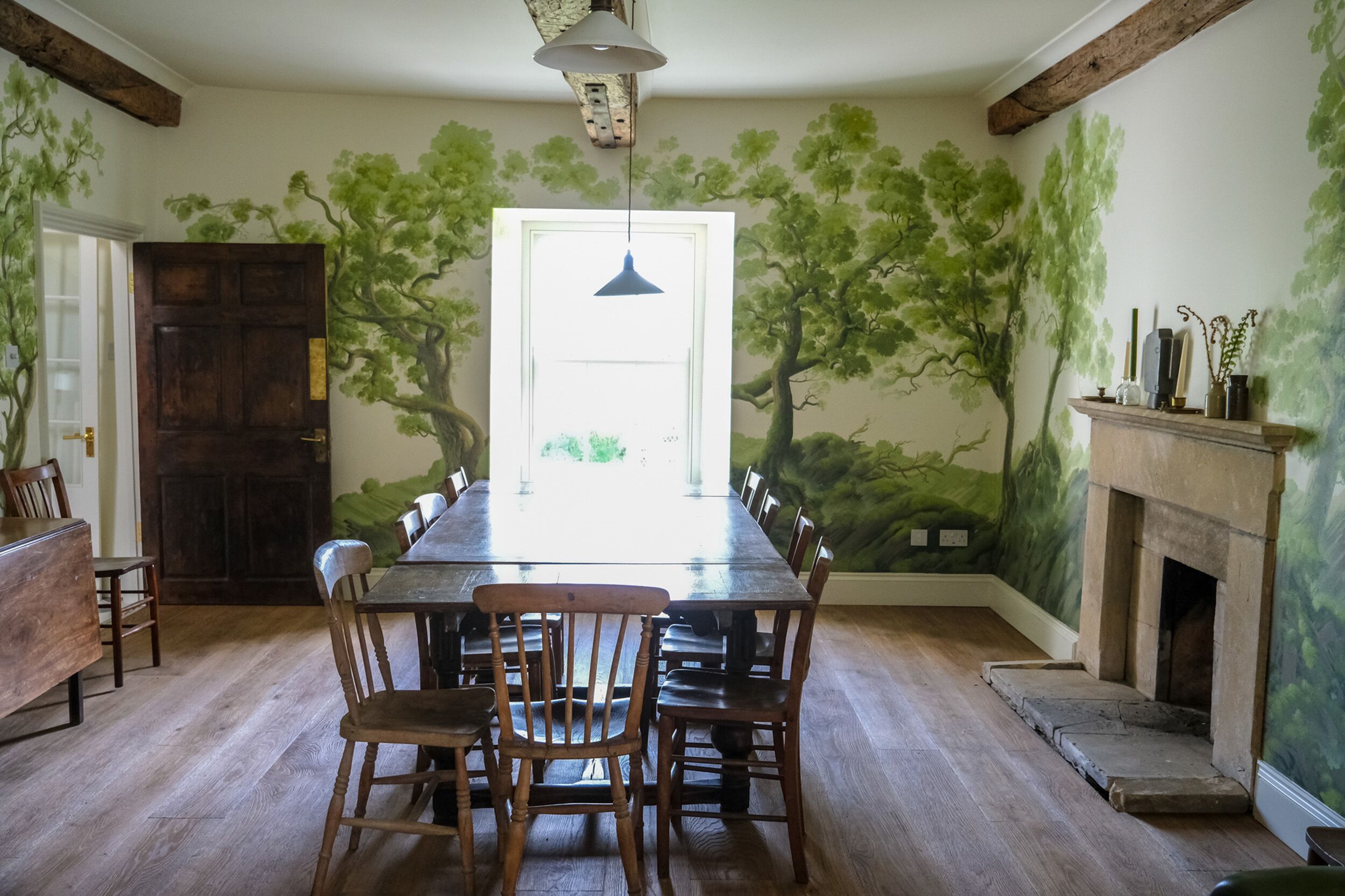 A dining room with a long wooden table, chairs, a fireplace, and a mural of trees on the wall.