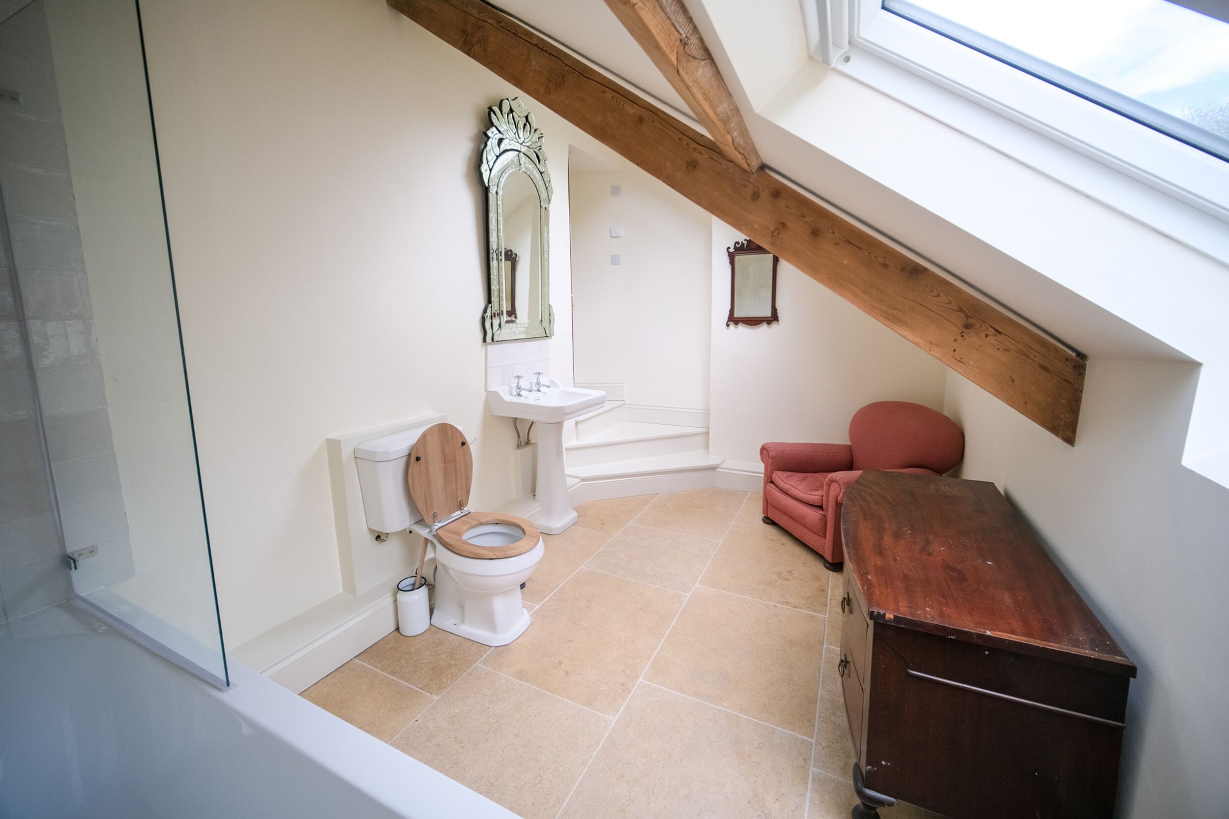 A small, compact bathroom with a wooden toilet, a white sink, and a red chair.