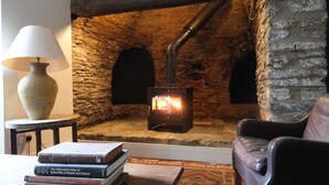 A stone fireplace with a wood-burning stove, a lamp, and books on a table in a cozy room. - Henllys (Crymych)