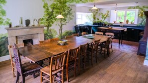 A dining area with a large wooden table and chairs, a fireplace, and a kitchen area with a window and plants. - Henllys (Crymych)