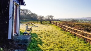 A rustic cabin with a wooden deck and picnic table, surrounded by a fenced yard and a view of rolling hills. - Henllys Lodge (Crymych)