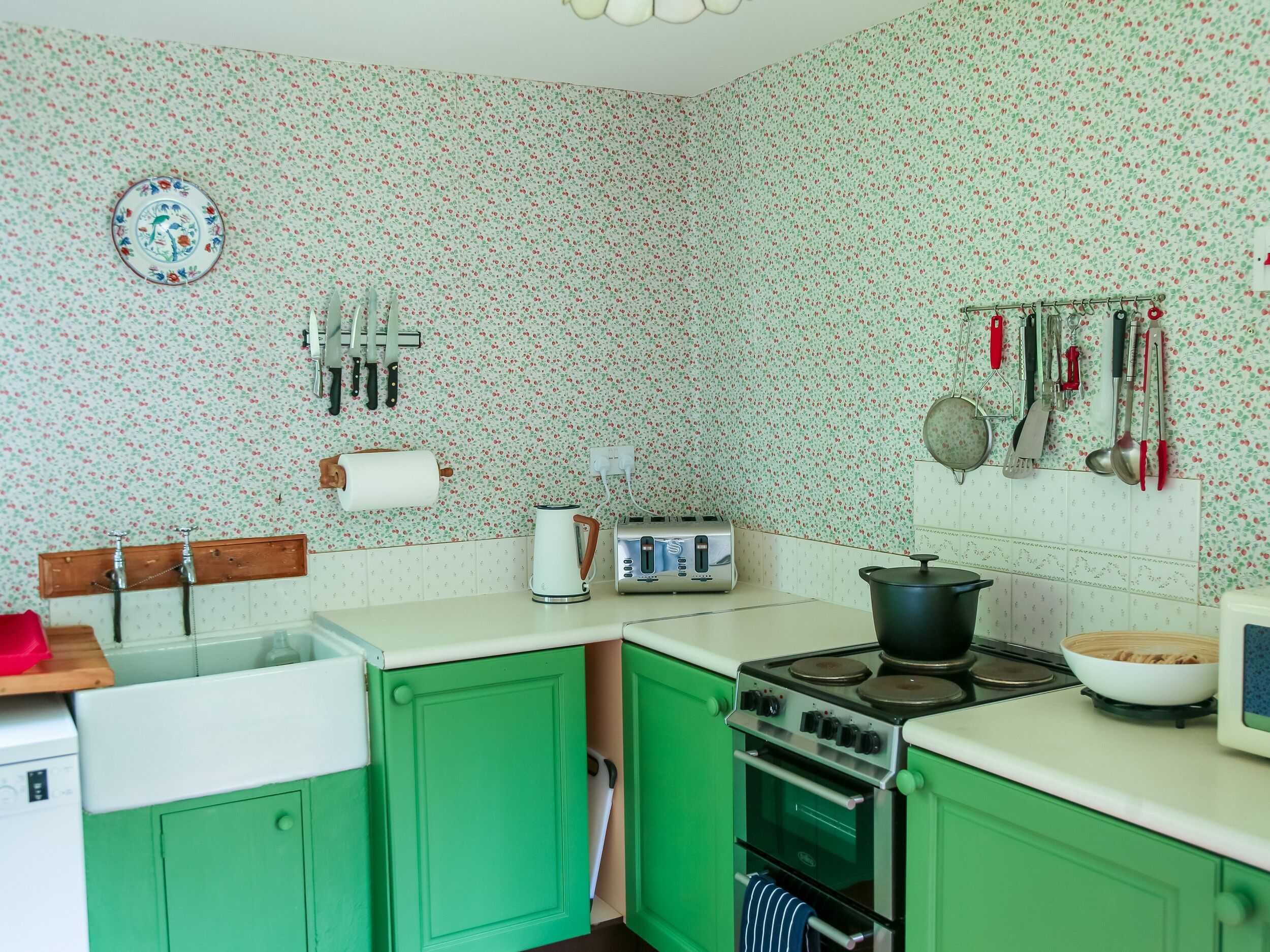 A kitchen with green cabinets, a stove, a kettle, and various utensils hanging on the wall.