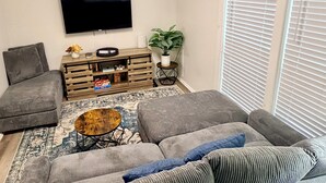 A living room with a grey sofa, a coffee table, a flat-screen TV, a wooden side table, and a potted plant.