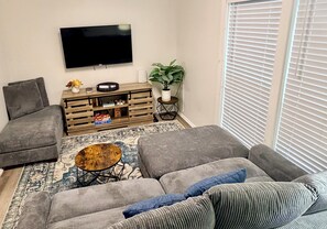 A living room with a grey sofa, a coffee table, a flat-screen TV, a wooden side table, and a potted plant.
