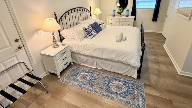 A bedroom with a bed, bedside tables, a nightstand, a mirror, a dresser, a window with curtains, and a patterned rug.