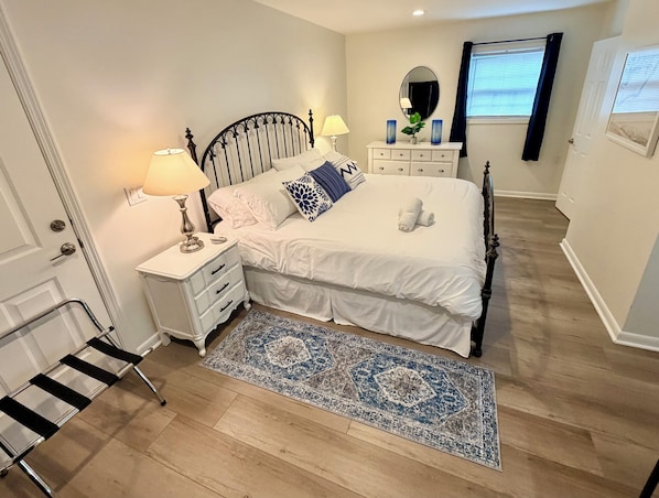 A bedroom with a bed, bedside tables, a nightstand, a mirror, a dresser, a window with curtains, and a patterned rug.