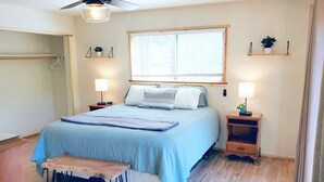 A bedroom with a bed, bedside tables, a nightstand, a bench, a ceiling fan, a window with blinds, and wall-mounted shelves with plants.