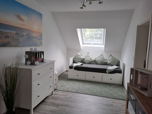 Ein Schlafzimmer mit geneigter Decke, einem Fenster mit Blick auf Grünflächen, einem Bett mit Kissen, einer Kommode und einem Gemälde an der Wand.