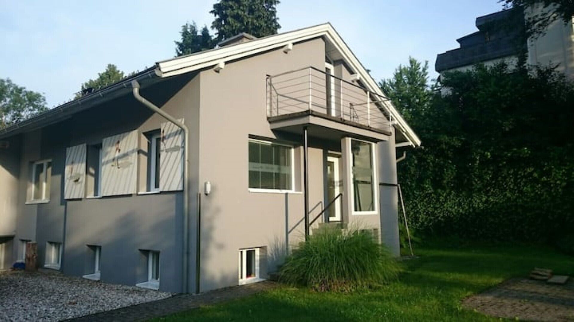 Ett modernt hus i två våningar med en balkong, stora fönster och en grusuppfart.