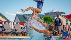 Ein Schwimmbad mit badenden Personen, einem Rettungsturm und Liegestühlen mit Flaggen.