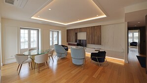 A modern living room with a glass dining table, light-colored chairs, and a TV.